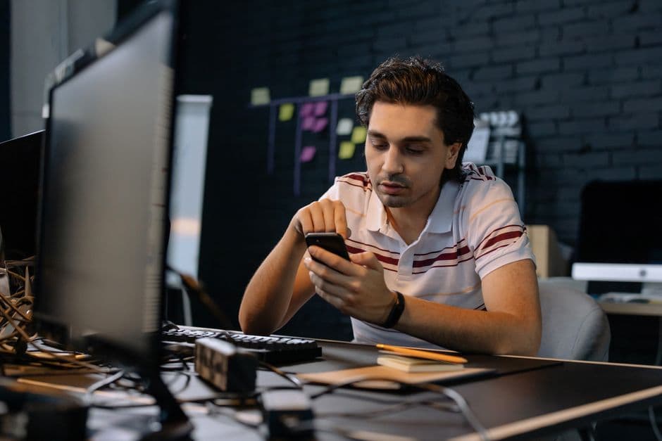 Young man in modern office using smartphone at his desk, focusing on multitasking work.